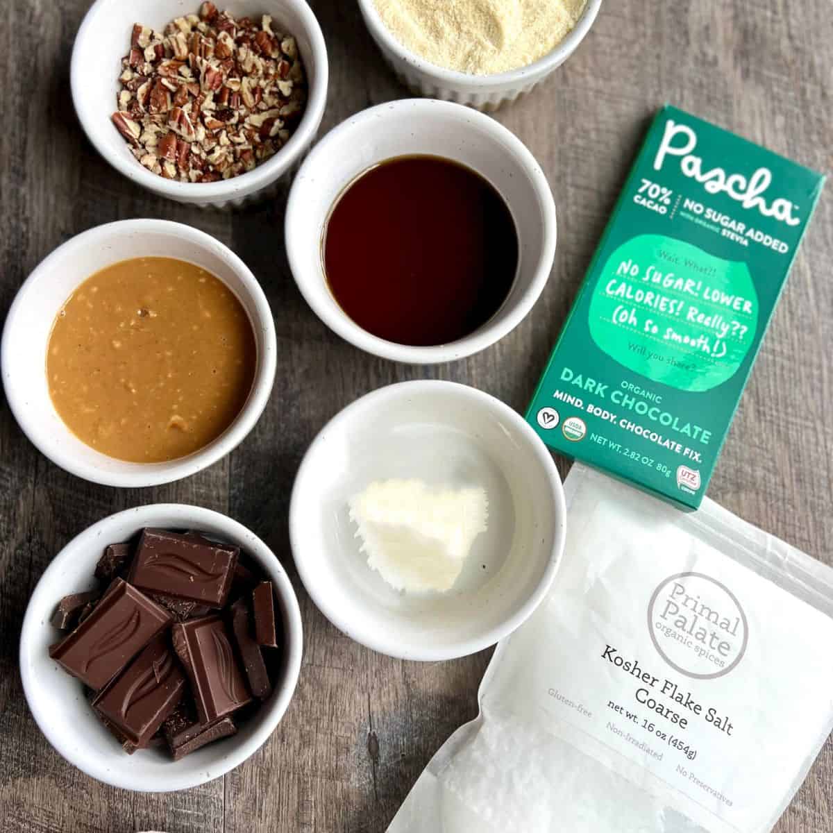 Ingredients - including chopped pecans, almond flour, almond butter, maple syrup and organic coconut oil laid out together in small white bowls with a chocolate bar and coarse kosher flake salt in a bag.