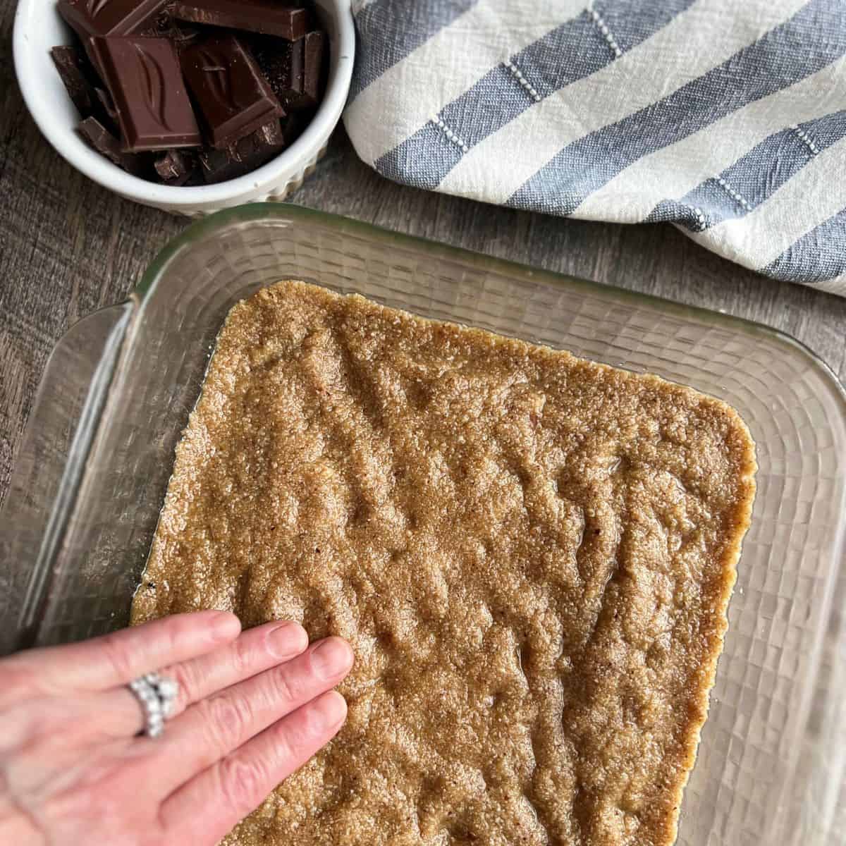 Using hands to press the base of the bars into an 8x8 glass pan with a small white bowl of chocolate squares to the side.