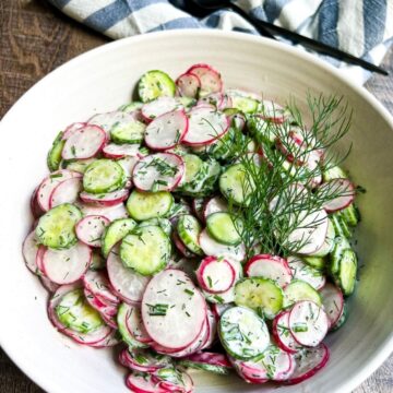 White serving bowl filled with crisp cucumber radish salad in a creamy dill dressing garnished with fresh dill.