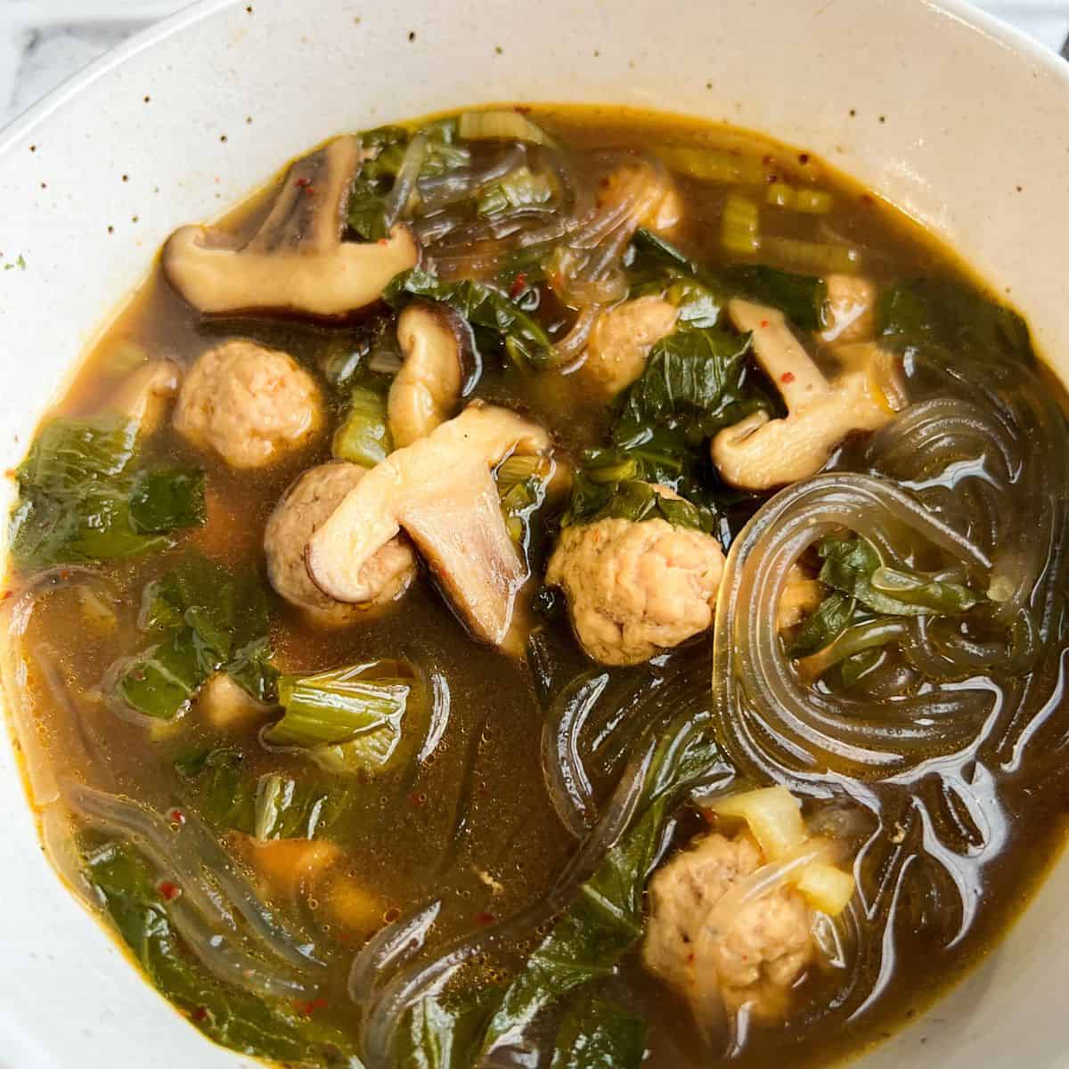 A close up of a speckled bowl filled with glass noodle ramen with ground pork meatballs, shiitake mushrooms, baby bok choy and green onions in a lemongrass ginger beef bone broth.