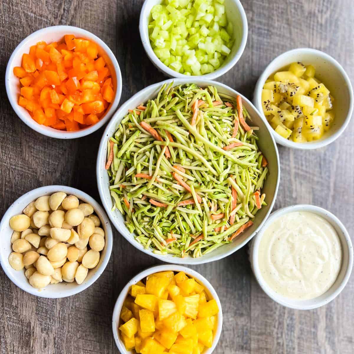 Ingredients for summer slaw laid out in white bowls, including broccoli slaw, avocado oil mayo, diced kiwi, celery, orange bell pepper, macadamia nuts and pineapple before everything is mixed together.