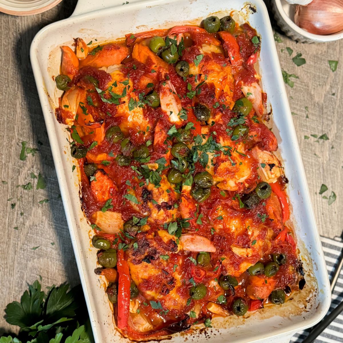 A baked casserole dish filled with cooked chicken, onions, red bell peppers, green olives, tomato sauce, and garnished with chopped parsley, placed on a wooden surface.