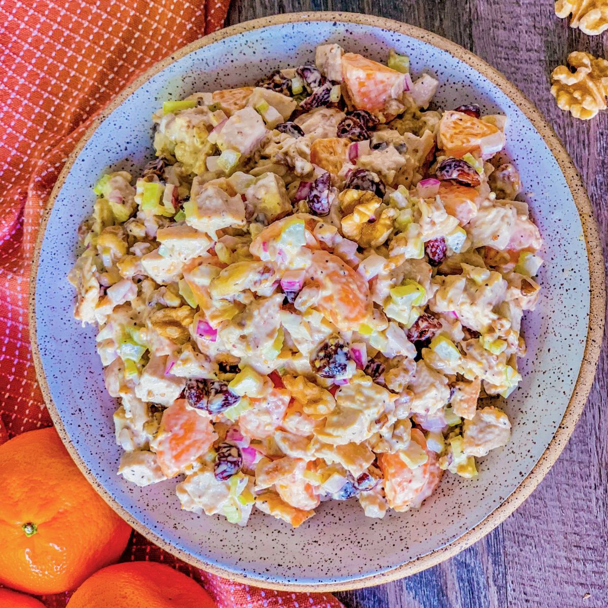 A bowl of creamy salad with chunks of apple, orange, celery, walnuts, and raisins, set on a wooden table next to whole oranges and a textured orange cloth.