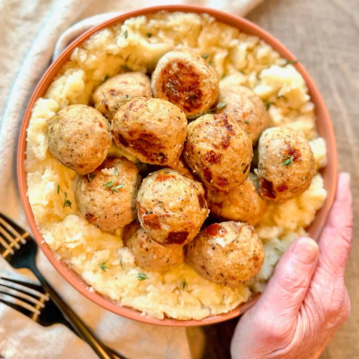 A bowl of mashed potatoes topped with several browned meatballs, garnished with herbs. A hand is holding the bowl, and black forks are visible on the side.