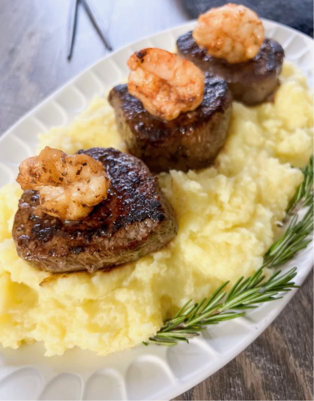 Three seared steak medallions topped with grilled shrimp, served on a bed of creamy mashed potatoes, garnished with a sprig of rosemary on a white oval plate.