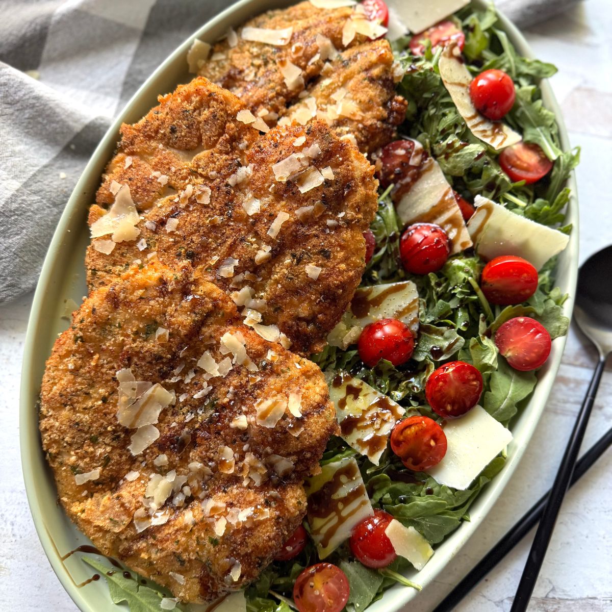 A plate with breaded, fried cutlets on one side and a fresh salad with arugula, cherry tomatoes, shaved cheese, and balsamic glaze on the other side, set on a light surface with a fork and spoon nearby.