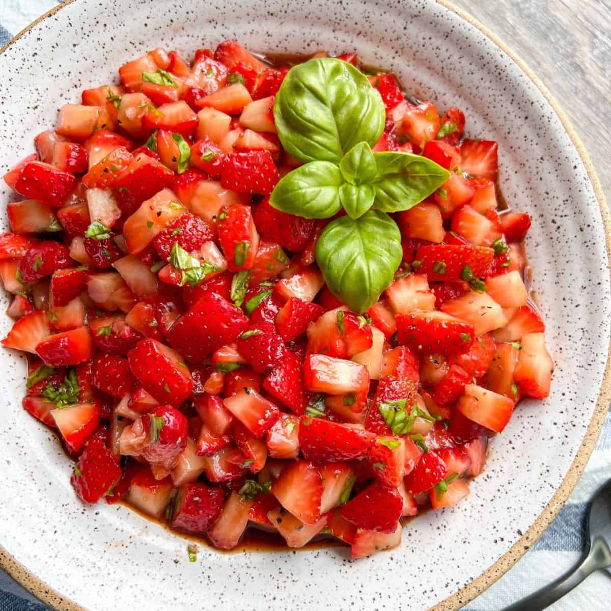 A white bowl filled with chopped fresh strawberries mixed with herbs, topped with a sprig of fresh basil for garnish.