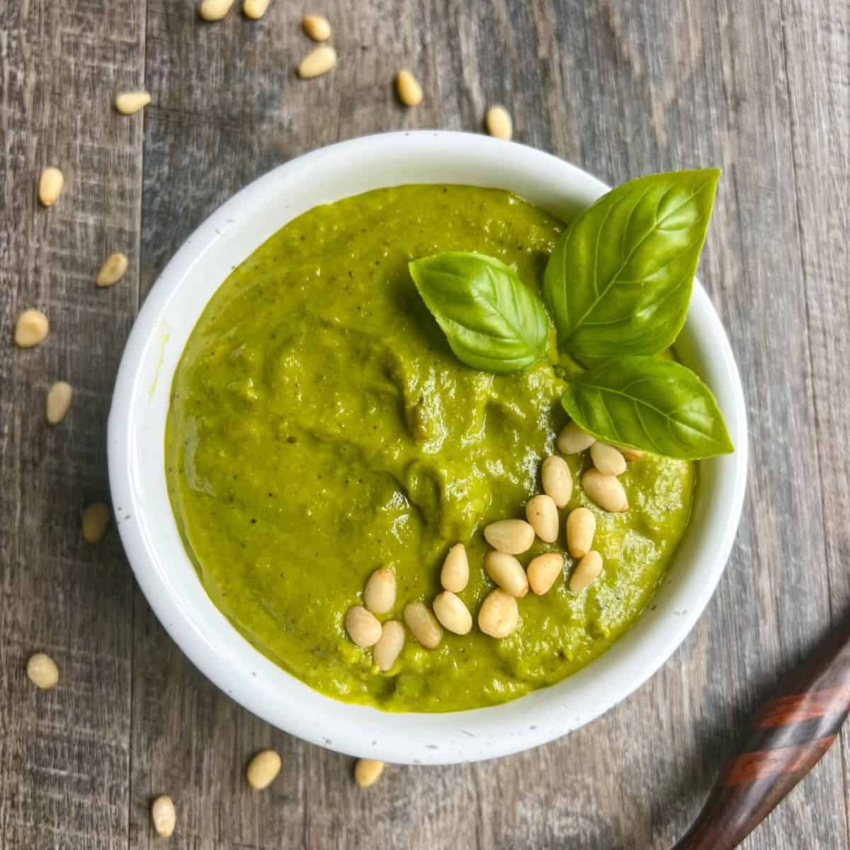 A white bowl of green pesto garnished with fresh basil leaves and pine nuts, placed on a rustic wooden surface. Some pine nuts are scattered around the bowl.