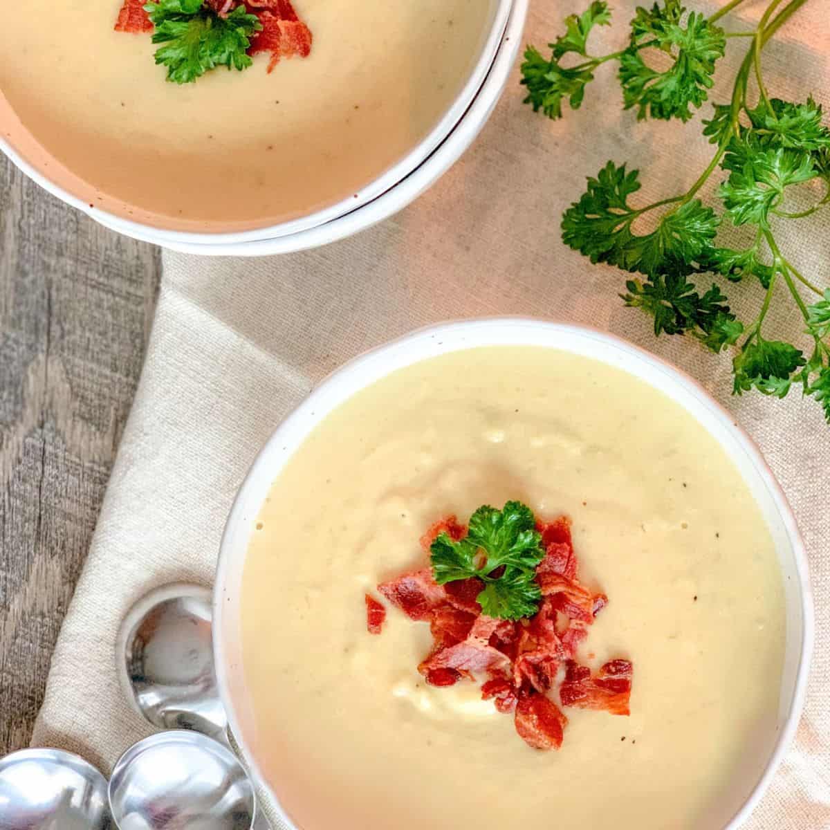 Two bowls of creamy soup topped with crumbled bacon and parsley sit on a beige cloth, with metal spoons and a sprig of fresh parsley nearby on a wooden surface.