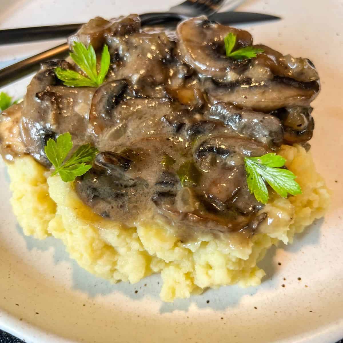 A serving of creamy mashed potatoes topped with a savory mushroom gravy, garnished with fresh parsley, on a white plate with utensils in the background.