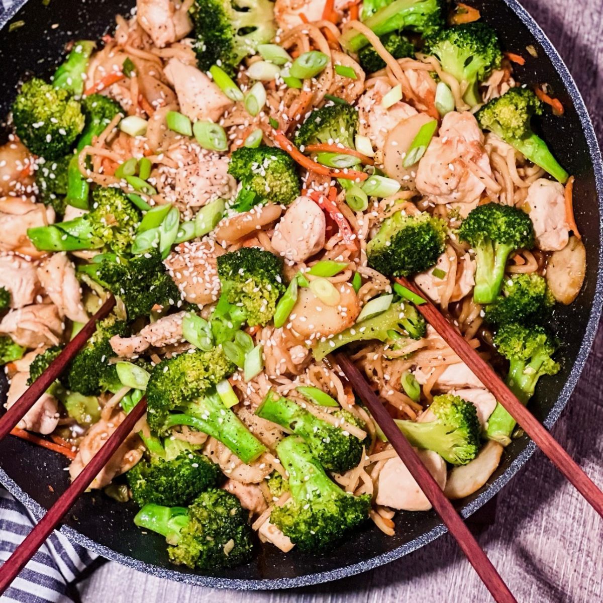 A skillet filled with chicken stir-fry, broccoli florets, sliced carrots, noodles, and green onions, topped with sesame seeds. Two pairs of chopsticks rest in the food, ready for serving.