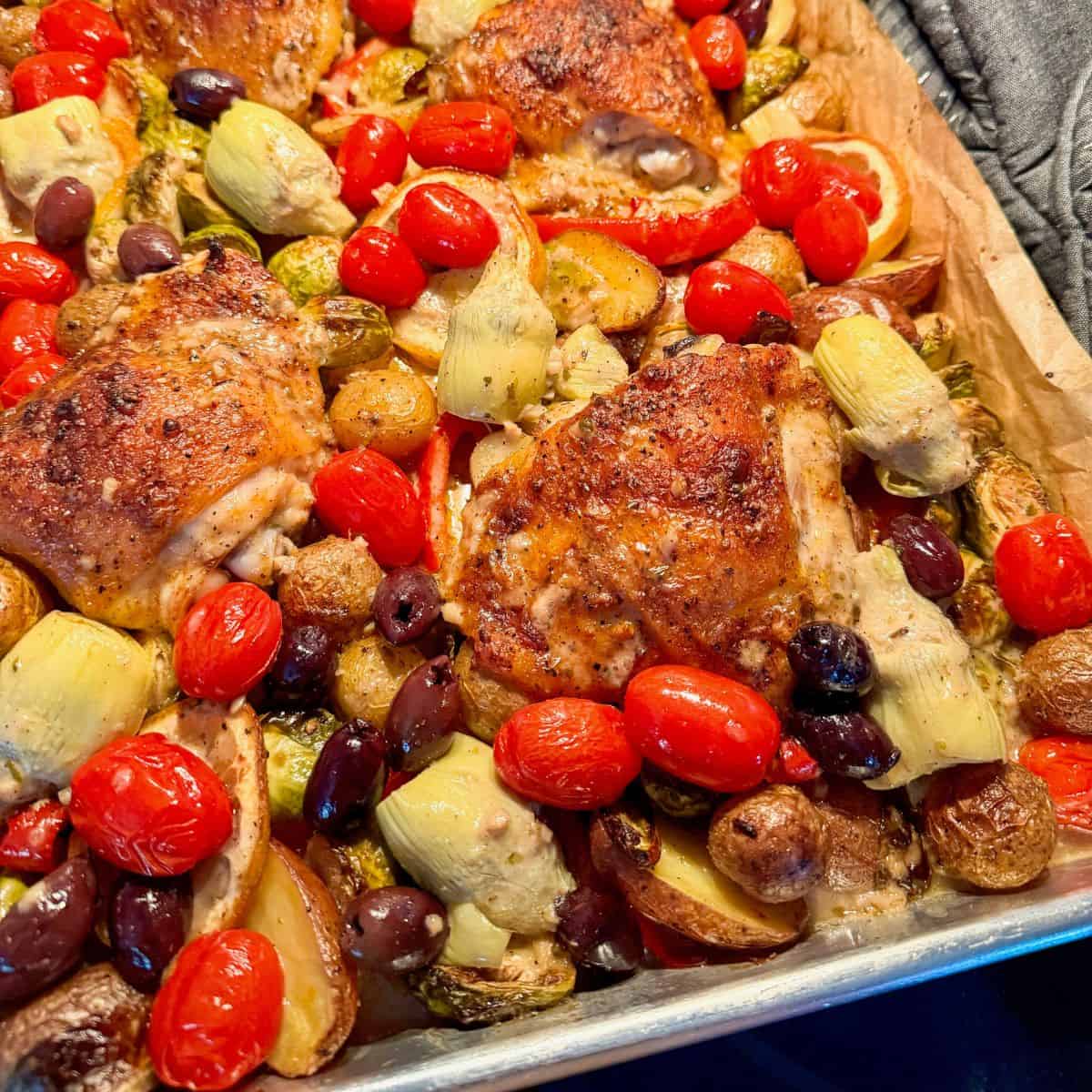 A sheet pan filled with roasted chicken thighs, cherry tomatoes, artichoke hearts, olives, Brussels sprouts, and halved baby potatoes, all seasoned and cooked together.