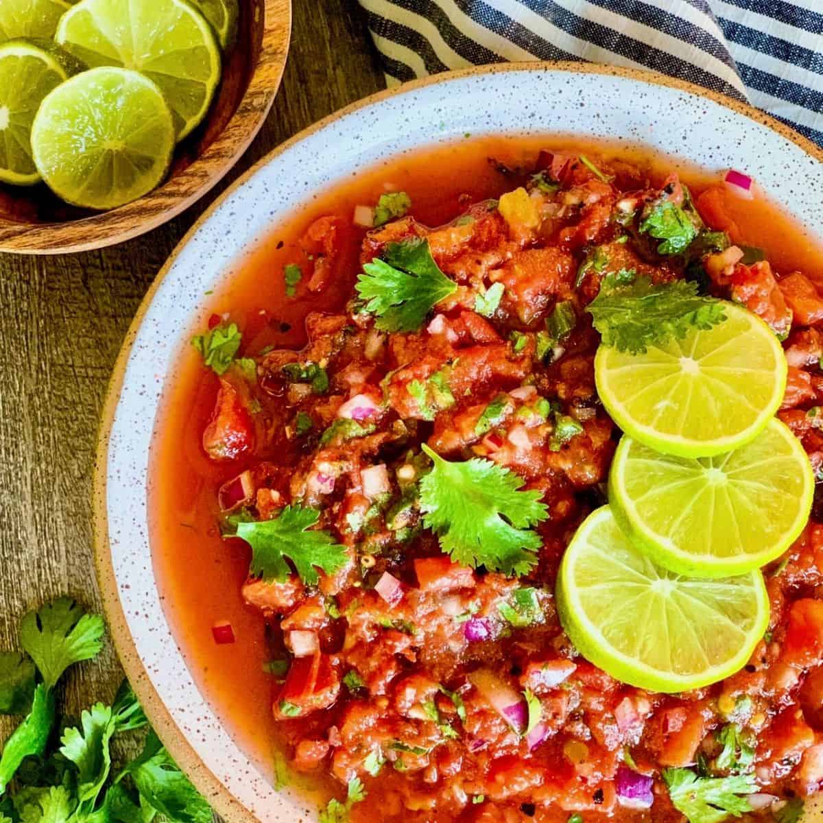 A bowl of fresh salsa made with diced tomatoes, onions, cilantro, and peppers, garnished with lime slices and cilantro leaves. A wooden bowl of lime wedges sits nearby on a striped cloth.