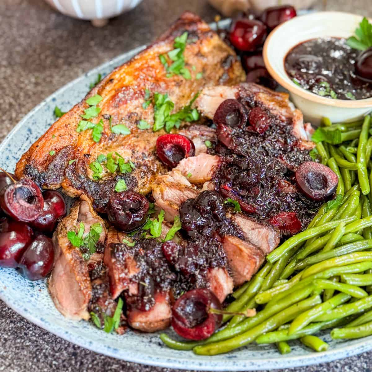 Sliced roast pork topped with cherry sauce and fresh cherries, garnished with parsley, served with a side of green beans on a blue and white plate. A small bowl of extra cherry sauce sits beside the meat.
