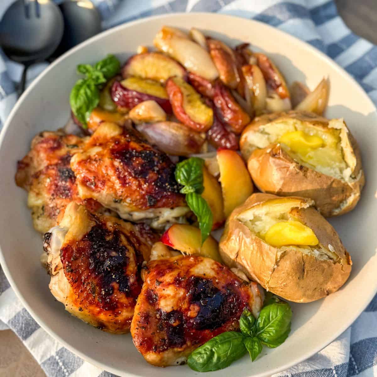 A plate with grilled chicken thighs, baked potatoes with butter, roasted onions and peaches, and a few fresh basil leaves, placed on a blue and white striped cloth.
