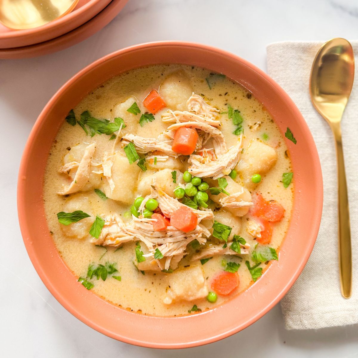 A bowl of creamy chicken and dumplings soup with shredded chicken, carrots, green peas, and parsley, served with a gold spoon and a white napkin on a marble surface.