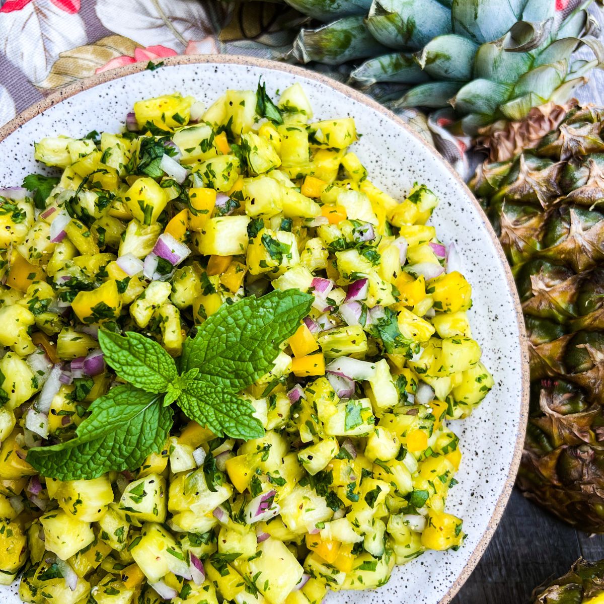 A bowl of fresh pineapple salsa with diced pineapple, red onion, cilantro, and yellow bell pepper, garnished with mint leaves. Whole pineapples and a floral napkin are visible in the background.