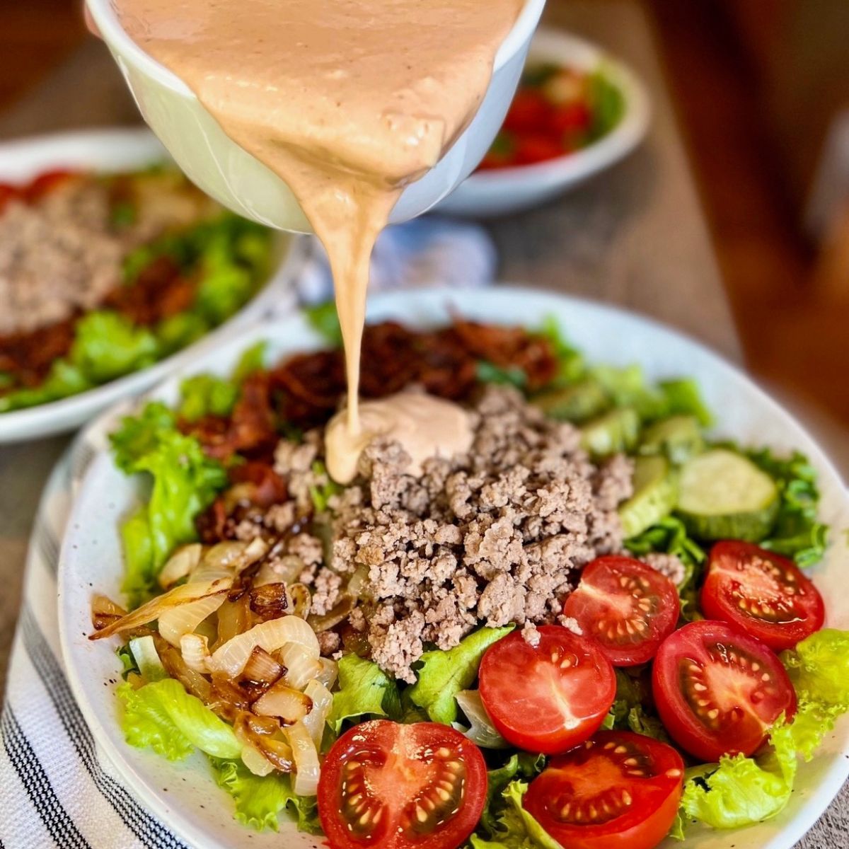 A bowl of creamy dressing is being poured over a salad with ground meat, lettuce, sliced tomatoes, cucumbers, and caramelized onions on a white plate. Another salad bowl is visible in the background.