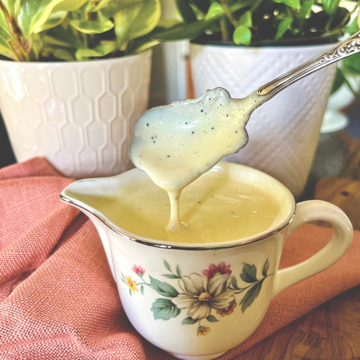 A spoon holds up thick, creamy sauce above a floral-patterned creamer jug. The scene is set on a pink cloth with white potted plants and green leaves in the background.