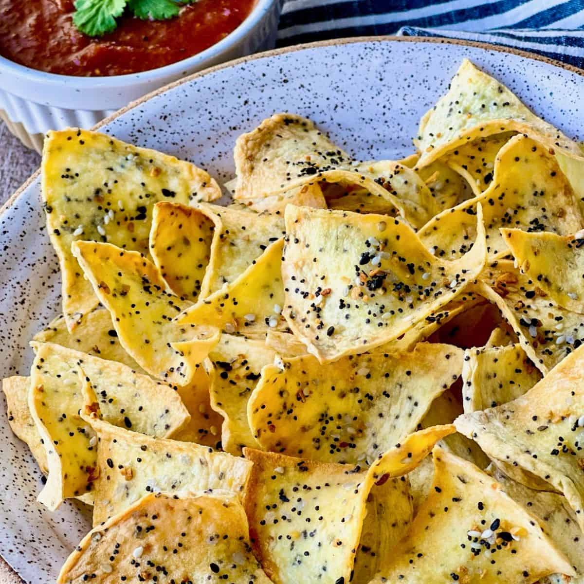 A plate of seasoned tortilla chips sprinkled with poppy and sesame seeds, served next to a bowl of red salsa.