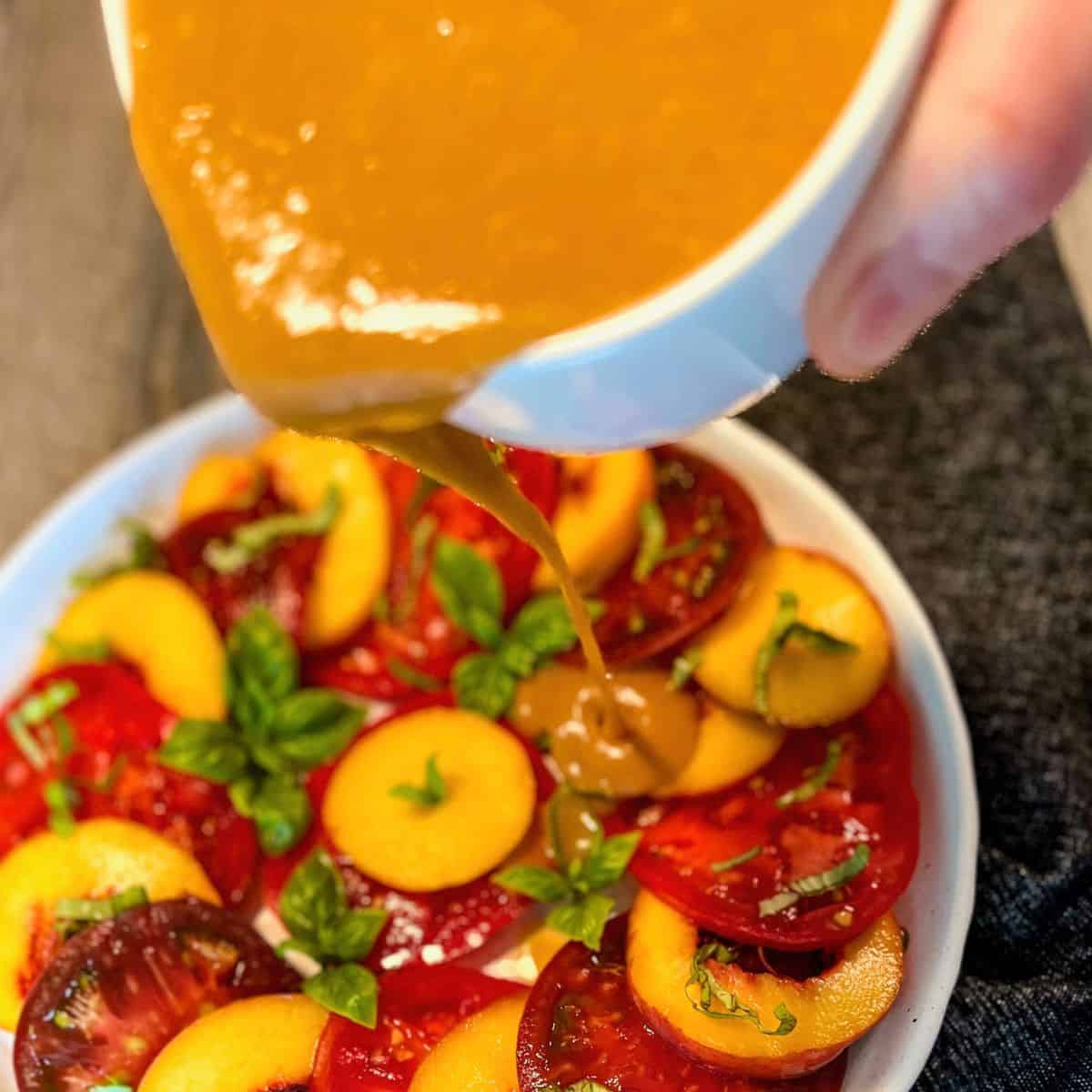 A hand pours a thick brown dressing over a salad made of sliced peaches, tomatoes, and fresh basil leaves arranged on a white plate.