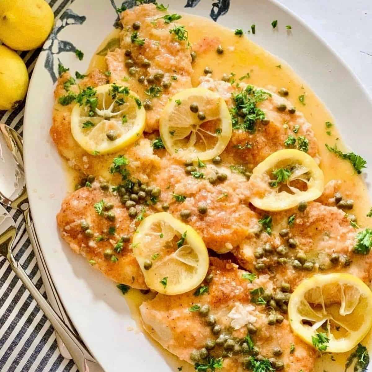 A platter of chicken piccata topped with lemon slices, capers, and chopped parsley in a light sauce, with whole lemons beside the plate and cutlery on a striped napkin.