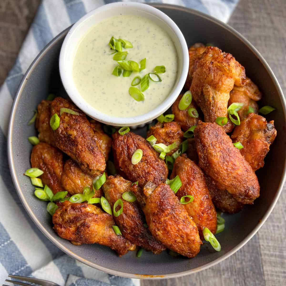 A bowl of crispy chicken wings garnished with sliced green onions, served with a small bowl of creamy dipping sauce on the side, placed on a striped cloth.