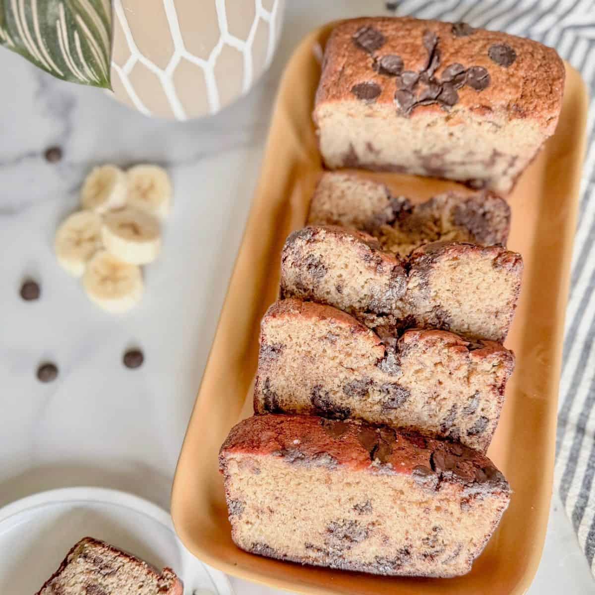 A rectangular plate holds four thick slices and one loaf of chocolate chip banana bread. Banana slices and chocolate chips are scattered nearby on a marble surface. A vase with green leaves is partially visible in the background.