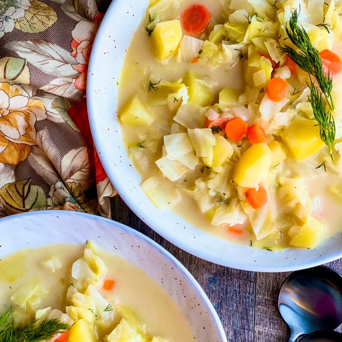 Two bowls of creamy vegetable soup with potatoes, carrots, cabbage, and fresh dill on a wooden table, next to a floral-patterned napkin and metal spoon.