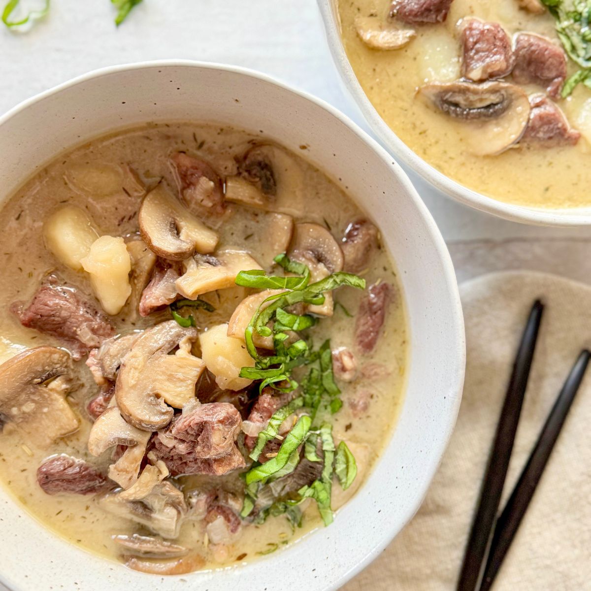 Two bowls of creamy soup with chunks of beef, mushrooms, and sliced greens on top, placed on a light surface with chopsticks and a beige napkin nearby.