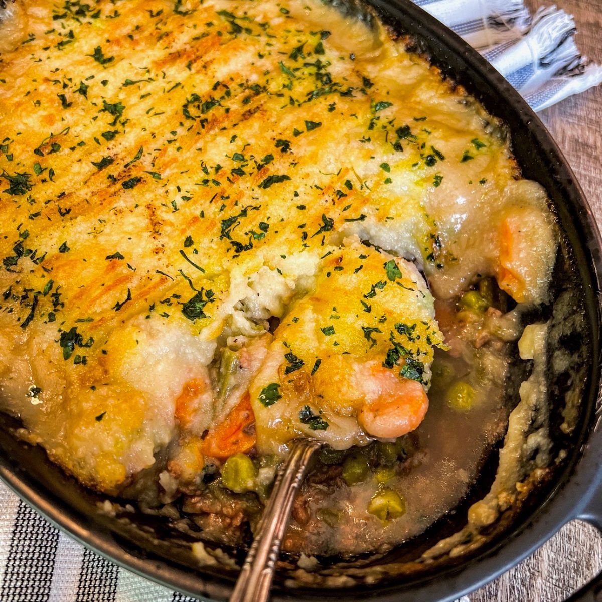 A close-up of a baked shepherd’s pie in a dish, topped with golden mashed potatoes and sprinkled with herbs. A spoonful has been taken out, revealing ground meat, peas, and carrots inside.