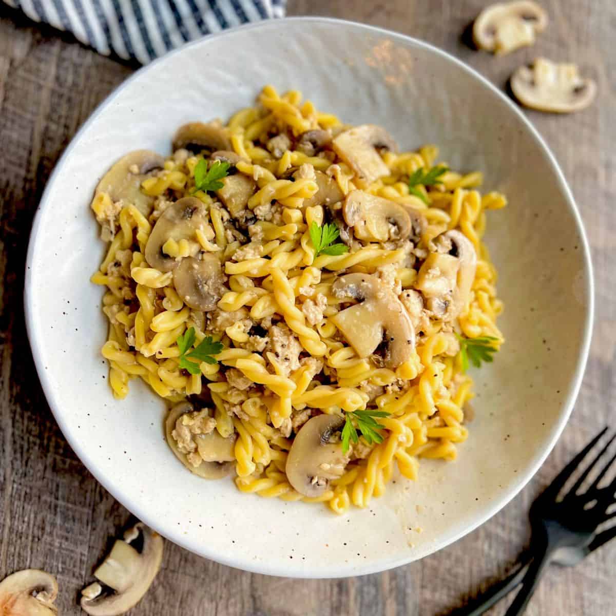 A white bowl filled with pasta spirals, sliced mushrooms, ground meat, and garnished with fresh parsley sits on a wooden table, with a striped napkin and scattered mushroom slices nearby.