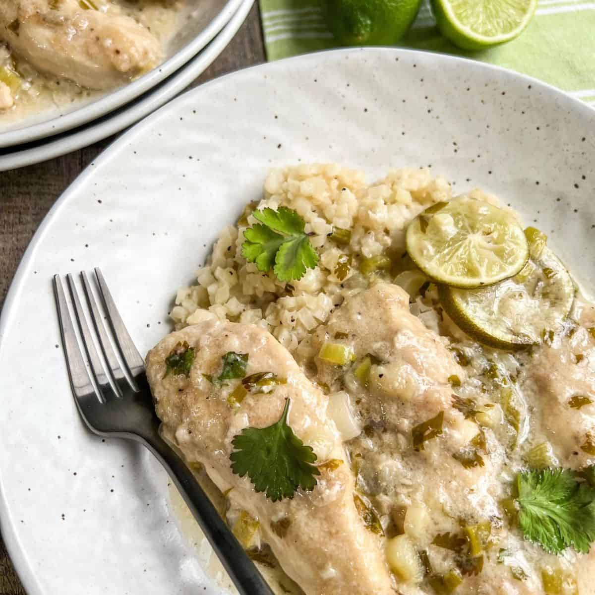 A white plate with cilantro lime chicken topped with lime slices and cilantro, served over rice. A fork rests on the side, and a lime wedge is visible in the background.