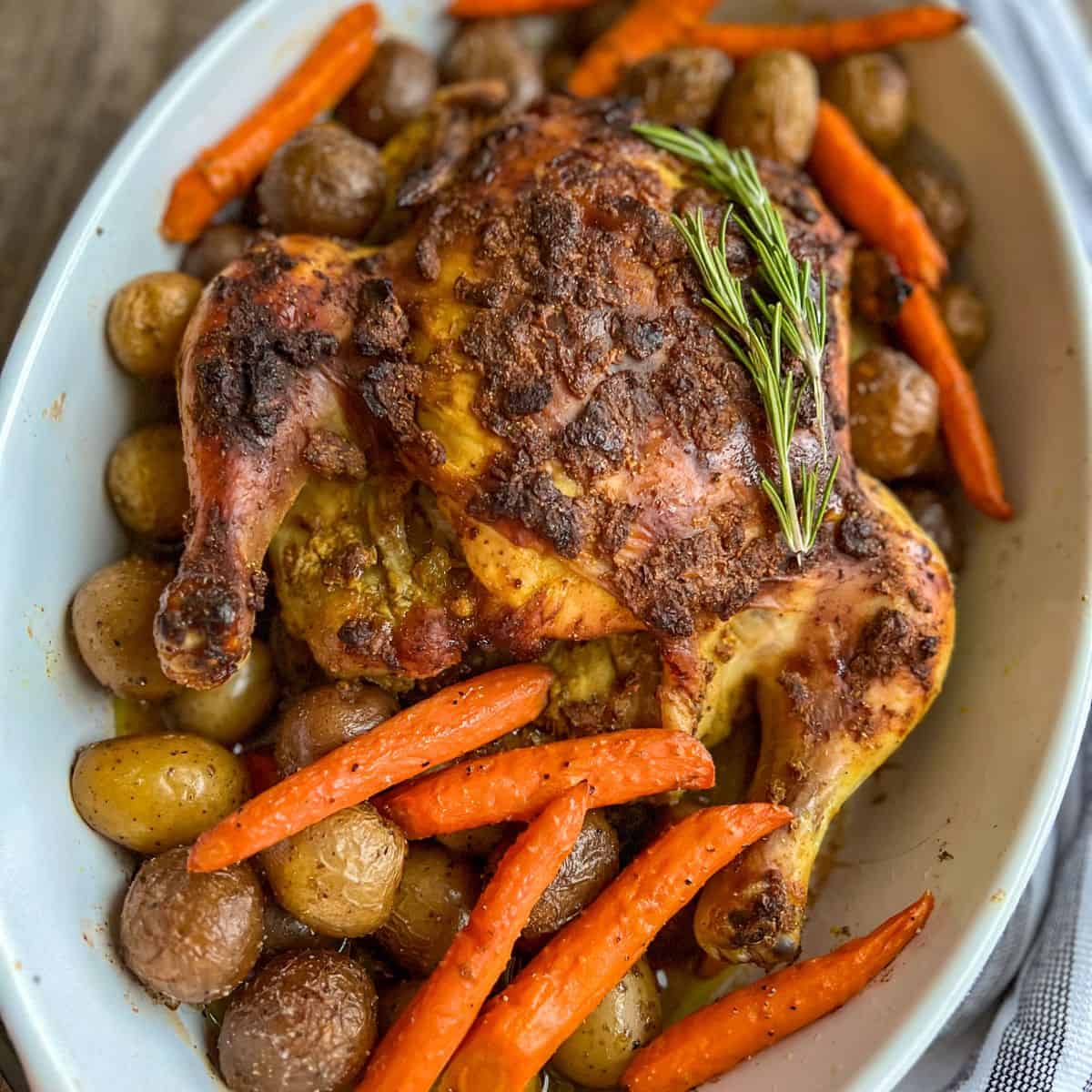 A roasted whole chicken garnished with a sprig of rosemary, surrounded by roasted baby potatoes and carrots in a white oval dish.