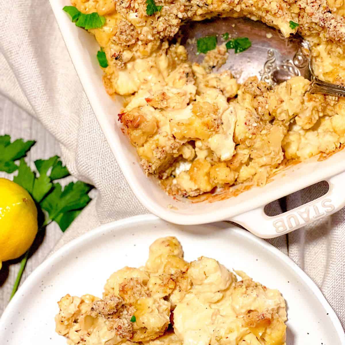 A baked cauliflower casserole with a golden, crumbly topping in a white dish, served with a spoon. A portion is on a plate nearby. Fresh parsley and lemons are placed beside the dish for garnish.