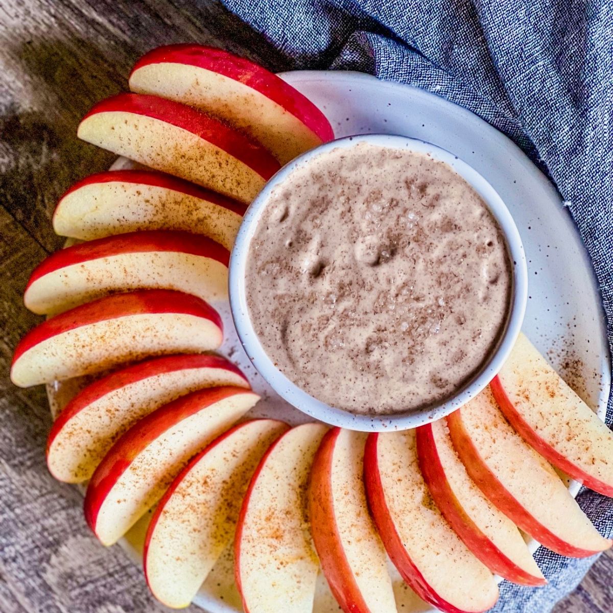 A plate of apple slices arranged in a circle around a bowl of creamy dip, all lightly sprinkled with cinnamon. A blue-gray cloth is partially visible in the background.