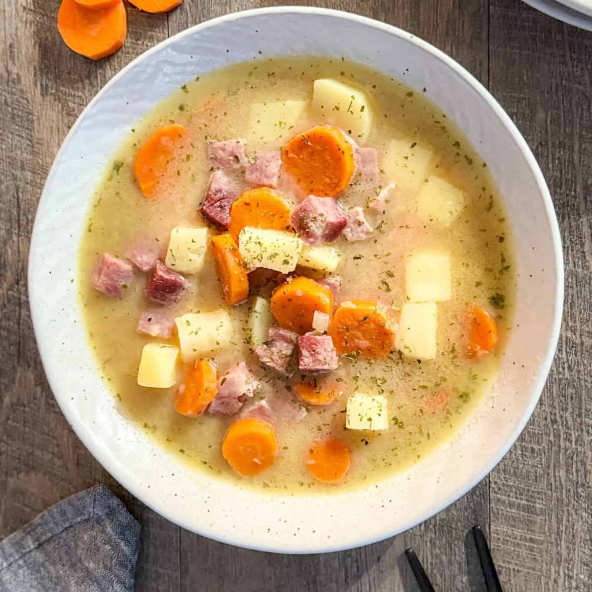 A bowl of hearty soup with chunks of ham, sliced carrots, and diced potatoes in a light broth, garnished with herbs, served on a wooden table.