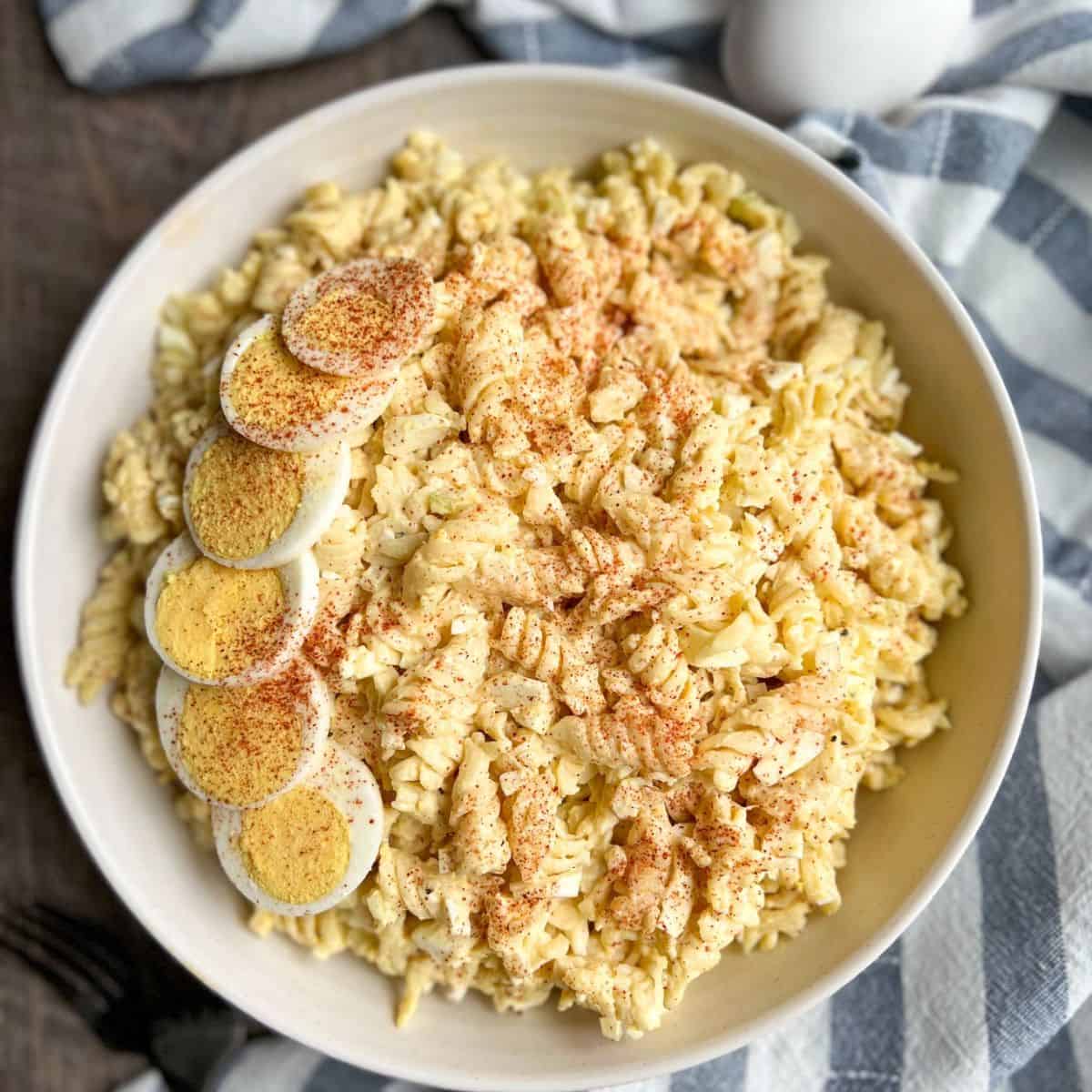 A bowl of creamy rotini pasta salad topped with sliced hard-boiled eggs and sprinkled with paprika, set on a cloth with a blue-and-white striped pattern.