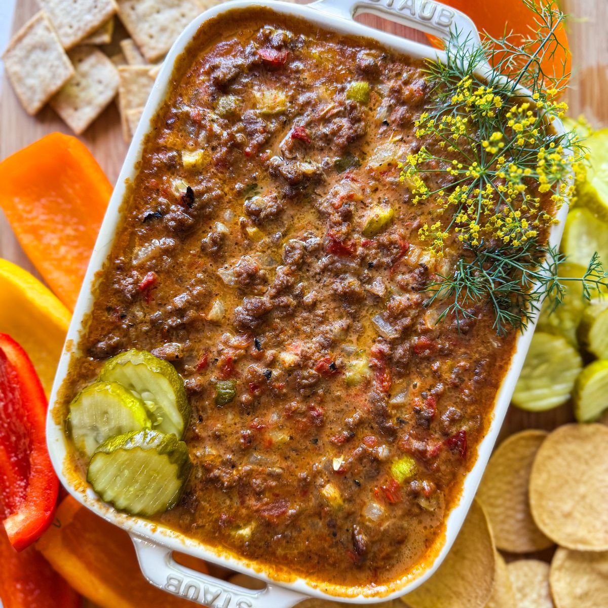 Rectangular dish filled with cheesy ground beef dip, topped with pickles and fresh dill. Surrounding the dish are crackers, sliced red and orange bell peppers, and pickles on a wooden board.
