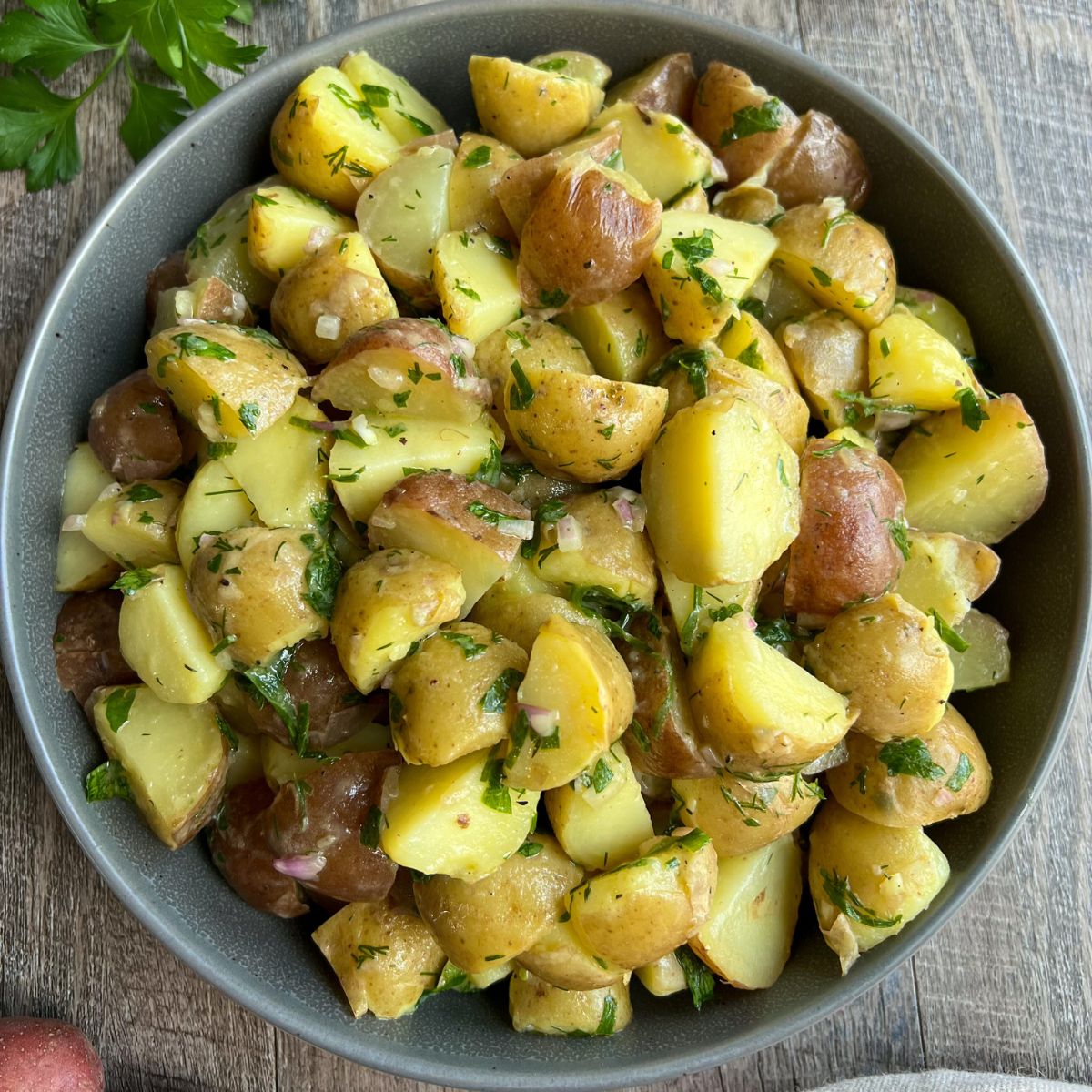 A bowl filled with chopped, cooked potatoes mixed with fresh herbs and seasonings, placed on a wooden surface. The potatoes have their skins on and are garnished with green herbs.