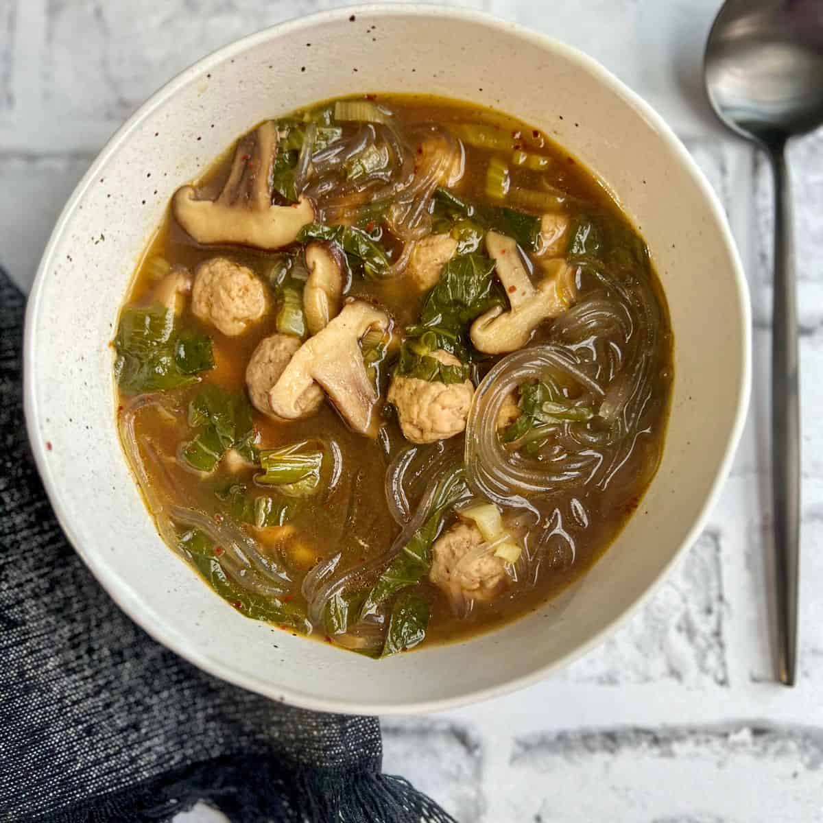 A bowl of soup with glass noodles, mushrooms, leafy greens, and pieces of meat in a clear brown broth, placed on a light brick surface next to a spoon and a dark textured cloth.