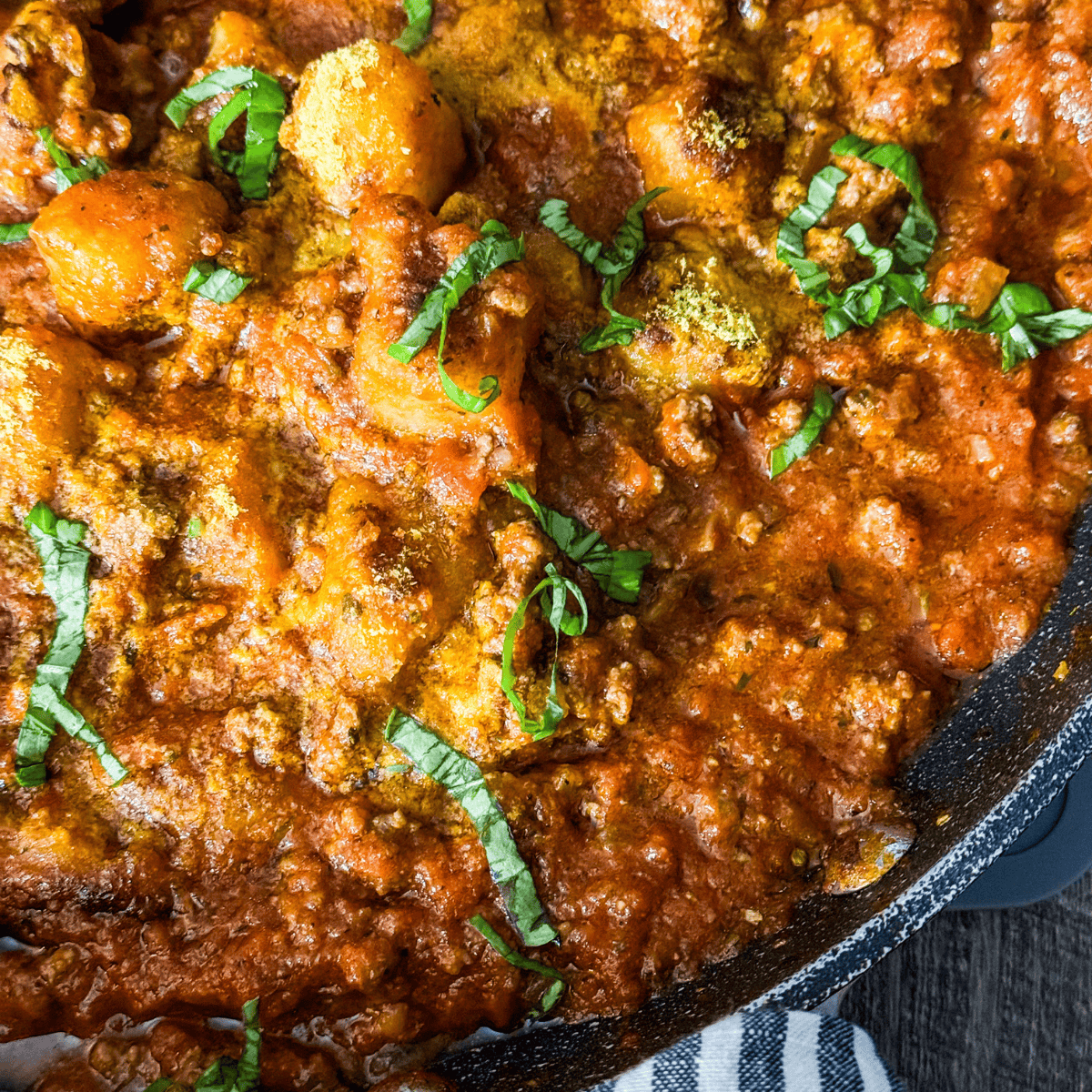 Close-up of a skillet filled with gnocchi in a rich, chunky tomato sauce, topped with chopped fresh basil and a sprinkle of seasoning, with a striped cloth partially visible underneath.