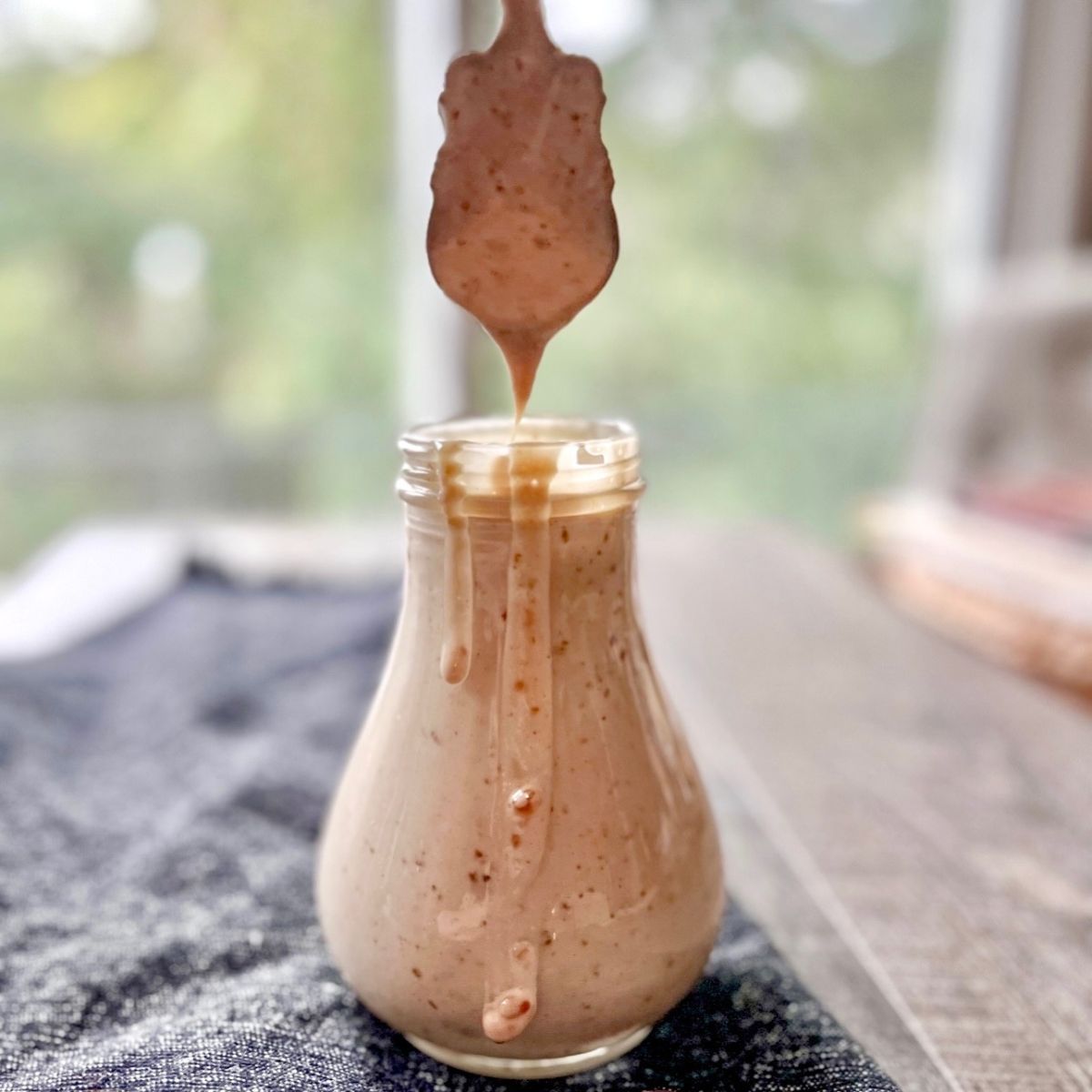 A glass jar filled with creamy, light brown sauce sits on a table, while a spoon held above drips some of the sauce back into the jar. The background is softly blurred.