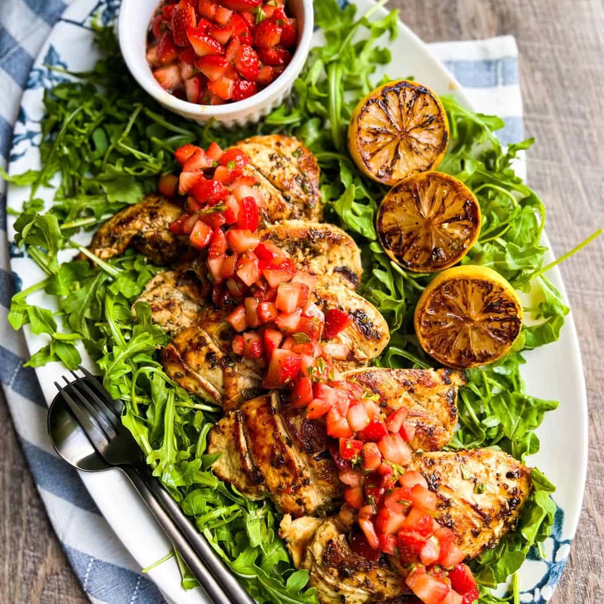 Grilled chicken breasts topped with fresh strawberry salsa, served on a bed of arugula with grilled lemon halves on a white platter; a small bowl of extra salsa sits nearby.