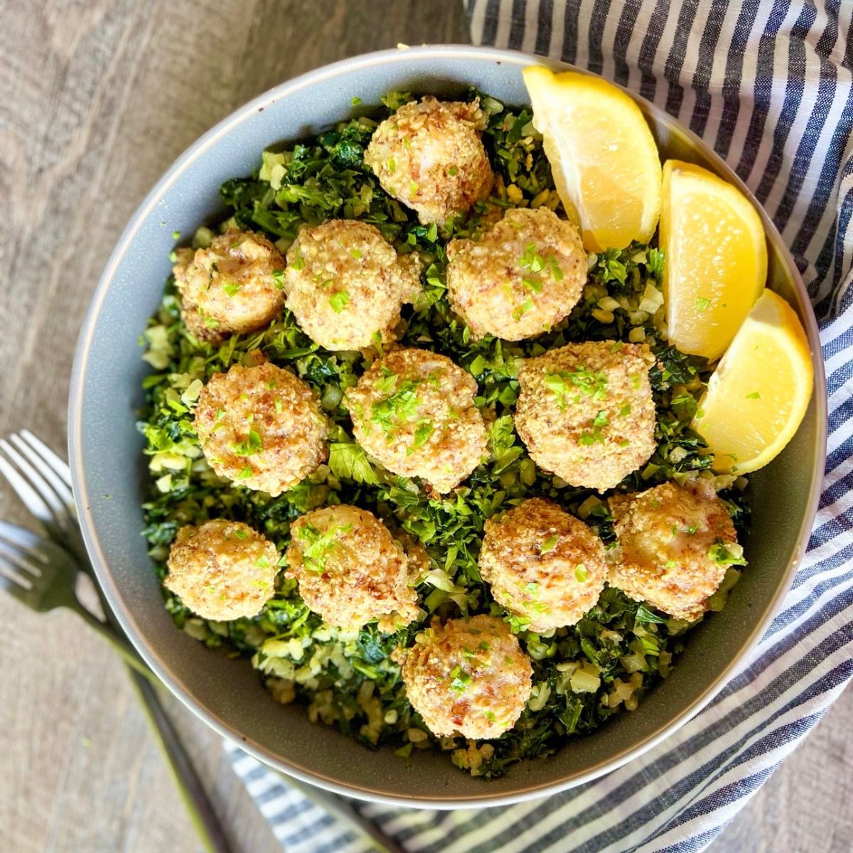 A bowl filled with breaded, baked shrimp arranged on a bed of chopped greens and cauliflower rice, garnished with fresh herbs and served with lemon wedges. A striped cloth and fork are beside the bowl.
