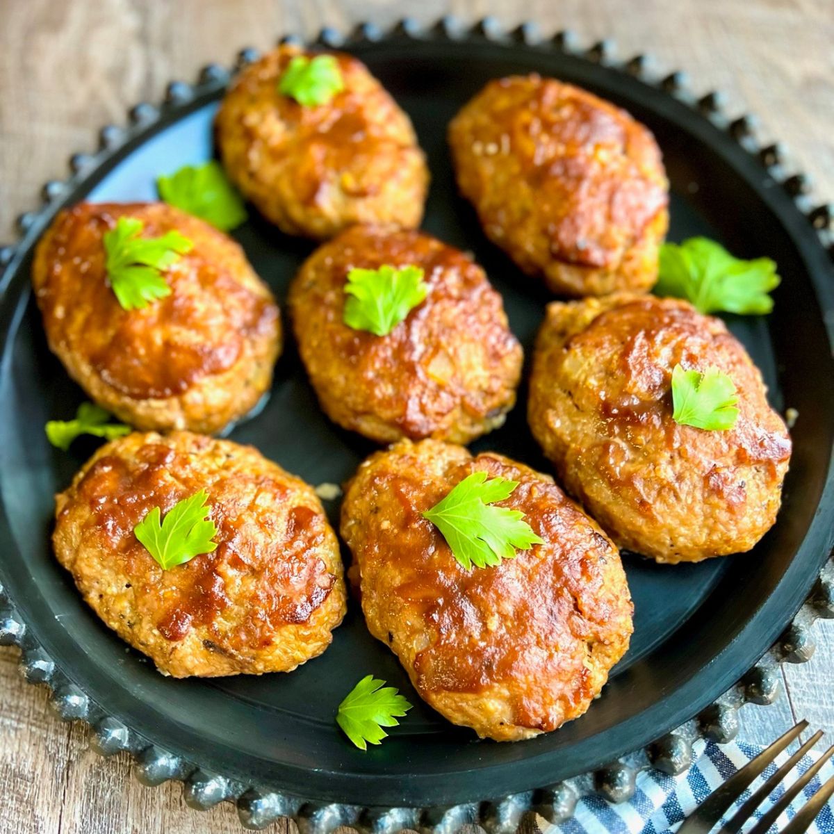 Eight oval-shaped meat patties garnished with fresh parsley leaves are arranged on a round black plate with a decorative edge, set on a wooden surface.