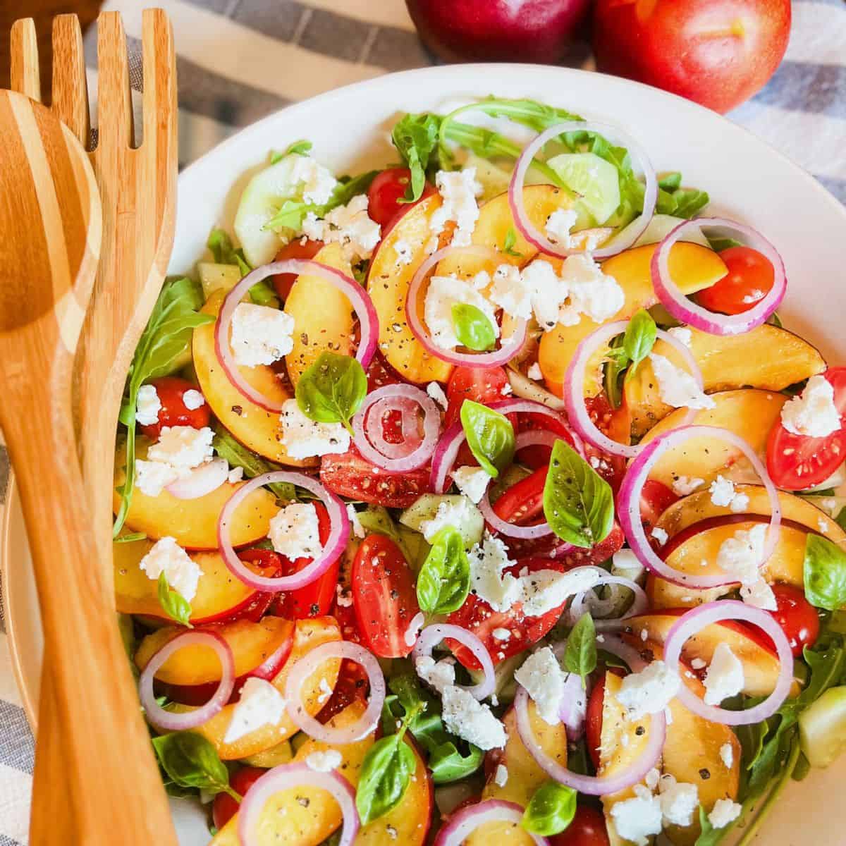 A fresh salad with sliced peaches, cherry tomatoes, red onion rings, feta cheese, and basil leaves on a bed of greens, served in a white bowl with wooden salad utensils nearby. Two whole peaches are in the background.