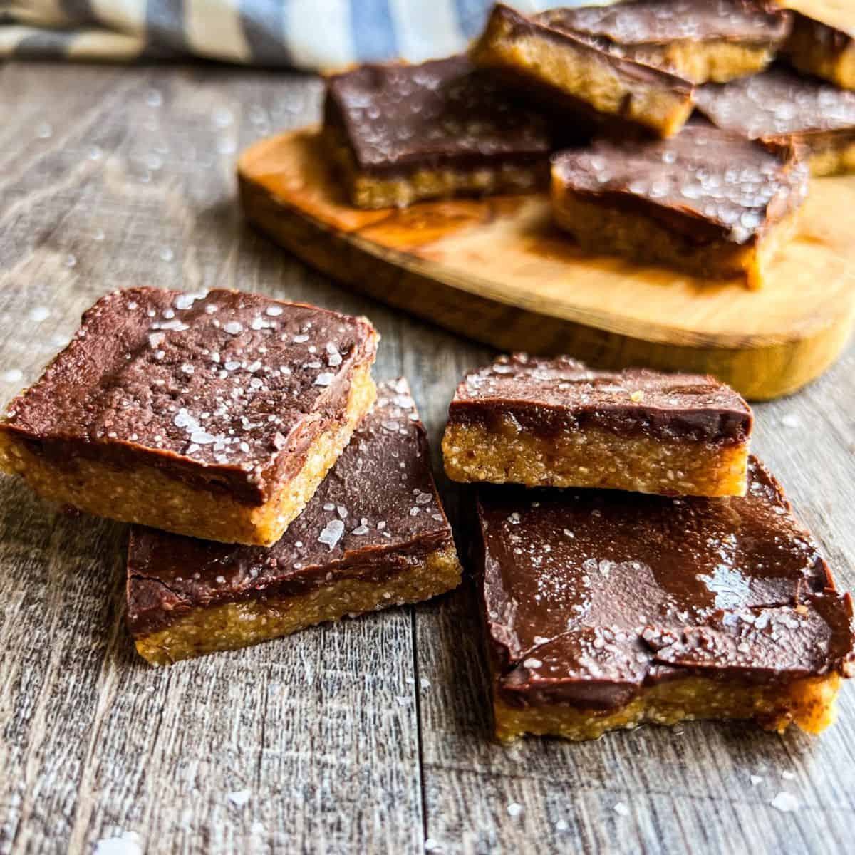 Four chocolate peanut butter bars with a sprinkle of sea salt are stacked and arranged on a wooden surface, with more bars on a wooden board in the background.