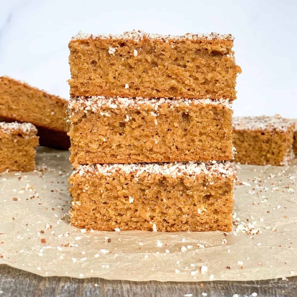 Three rectangular pieces of golden brown cake stacked on parchment paper, with a light dusting of shredded coconut or sugar on top. More cake pieces are visible in the background.