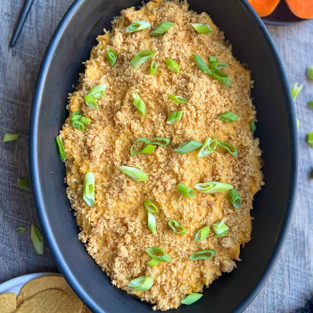 A black oval dish filled with a creamy, crumb-topped baked casserole, garnished with chopped green onions. The dish is set on a gray cloth with a small bowl of round crackers nearby.